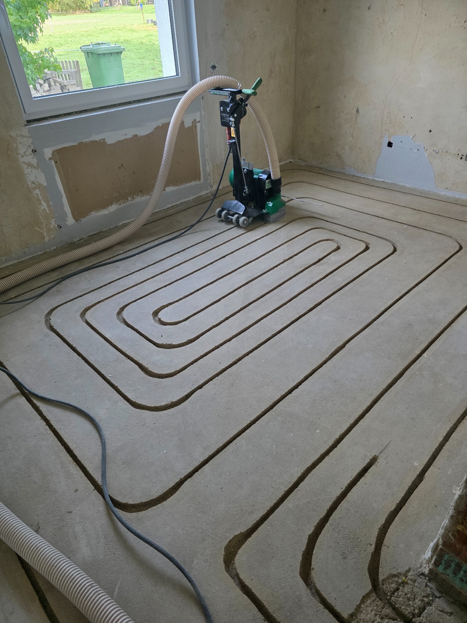 Fräsmaschine mit Absaugschlauch neben gefrästen Heizkanälen im vorhandenen Estrich
